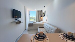 Una habitación de hotel moderna con televisión de pantalla plana, un sofá, una mesa de comedor con platos y tazas, y vistas al exterior.