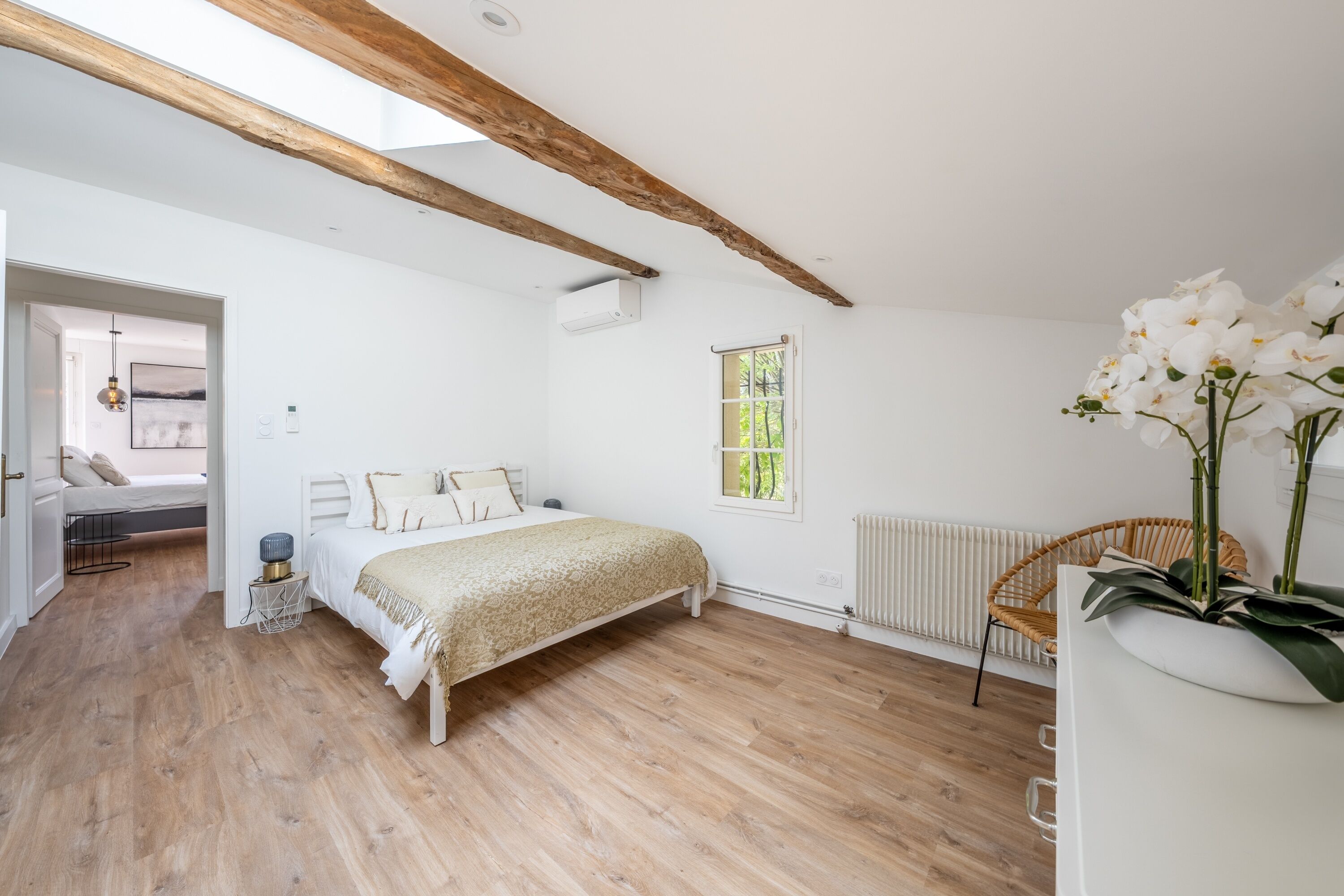 A bedroom with a wooden floor, a bed with a white headboard, a window with a view, and a wicker chair.