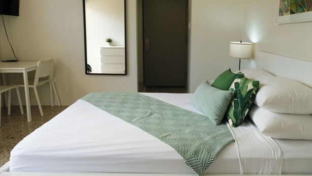 Quarto de hotel com cama, escrivaninha, cadeira, espelho, porta e abajur.