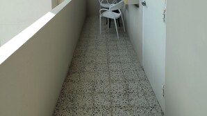 Um corredor estreito com chão de azulejos, paredes brancas e uma janela à esquerda. Há uma porta à direita, com um pequeno janelão em cima. Uma cadeira branca está encostada à parede, perto da porta.