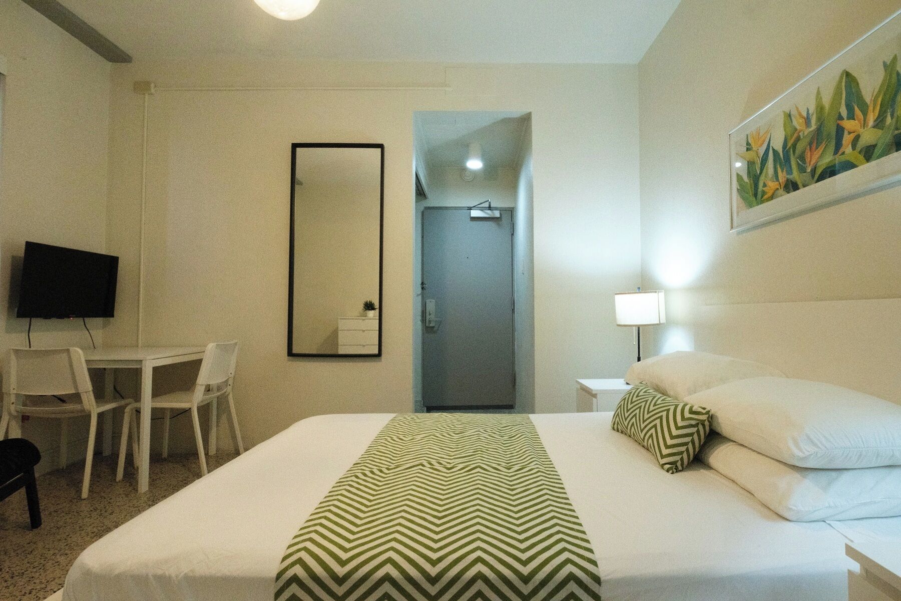 A hotel room with a bed, a desk with a chair, a television, a mirror, and a door.