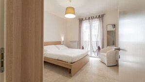 A modern bedroom with a wooden bed, a bedside lamp, a white chair, and a large window with curtains. - Alba di mare by Monholiday (Monopoli)