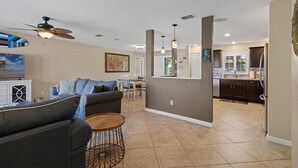 A modern living room with a sofa, coffee table, and ceiling fan. A dining area with chairs and a table is visible in the background. - Pelican's Cove (Port Orange)
