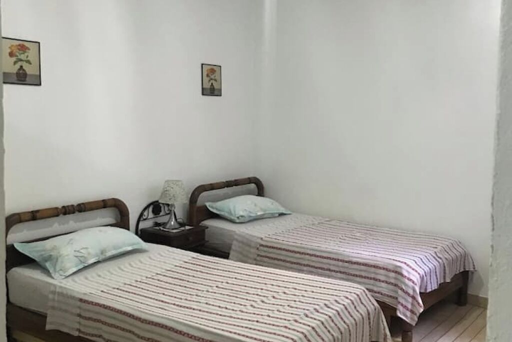 Een kleine, eenvoudige kamer met twee eenpersoonsbedden, elk met een houten hoofdbord en een gestreept dekbed. Er zijn twee nachtkastjes met lampen, en de muren zijn effen wit.