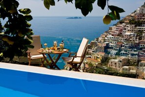A poolside view with a table and chairs overlooking a coastal town and the sea.