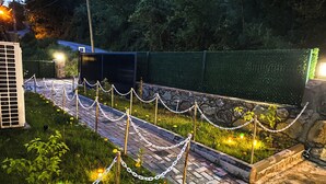 Un camino pavimentado con cerca de malla y luces decorativas, rodeado de follaje y un muro de piedra.