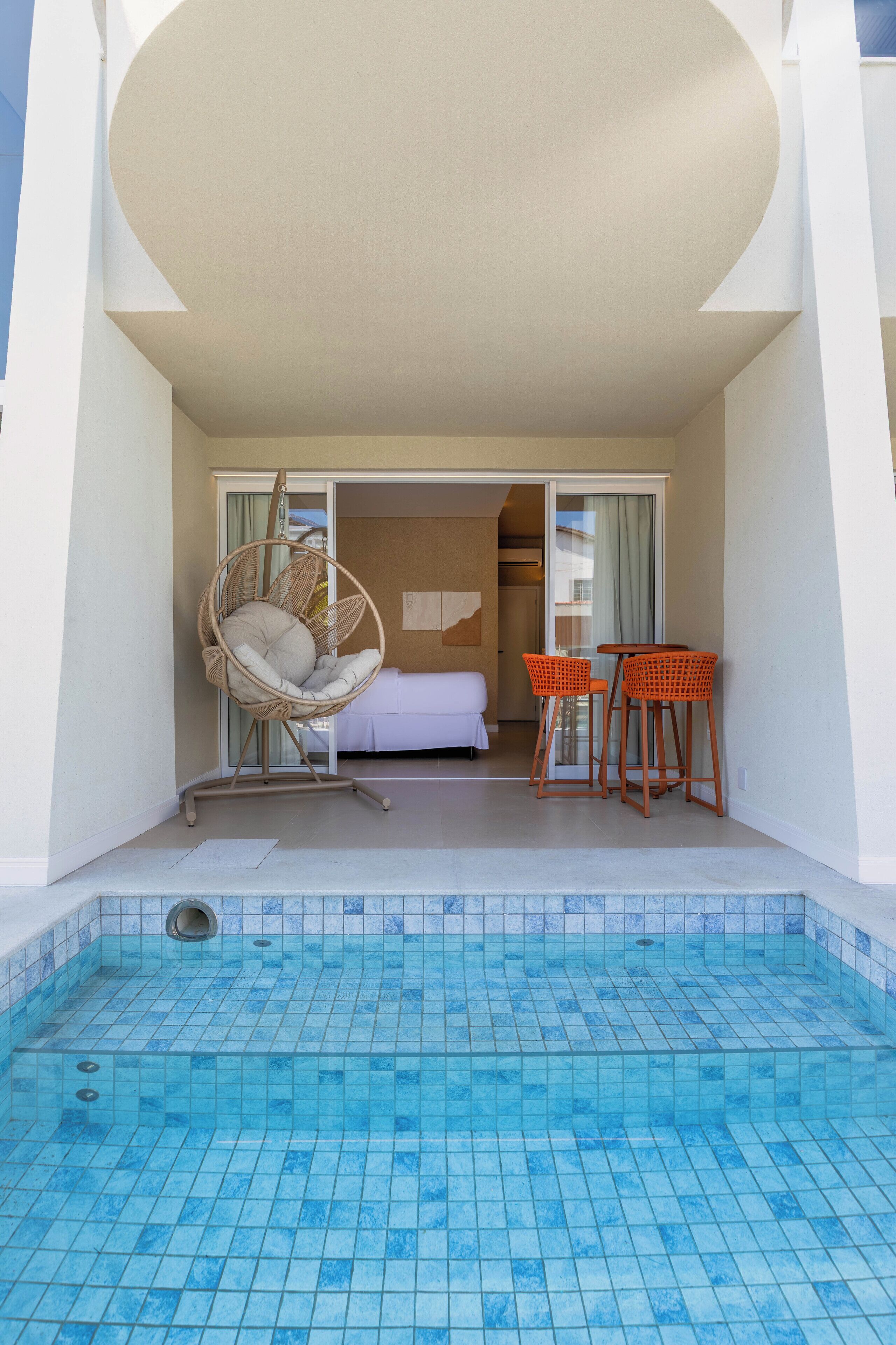 Un espace piscine agrémenté d’un fauteuil suspendu, de deux tabourets de bar et d’un lit en arrière-plan.
