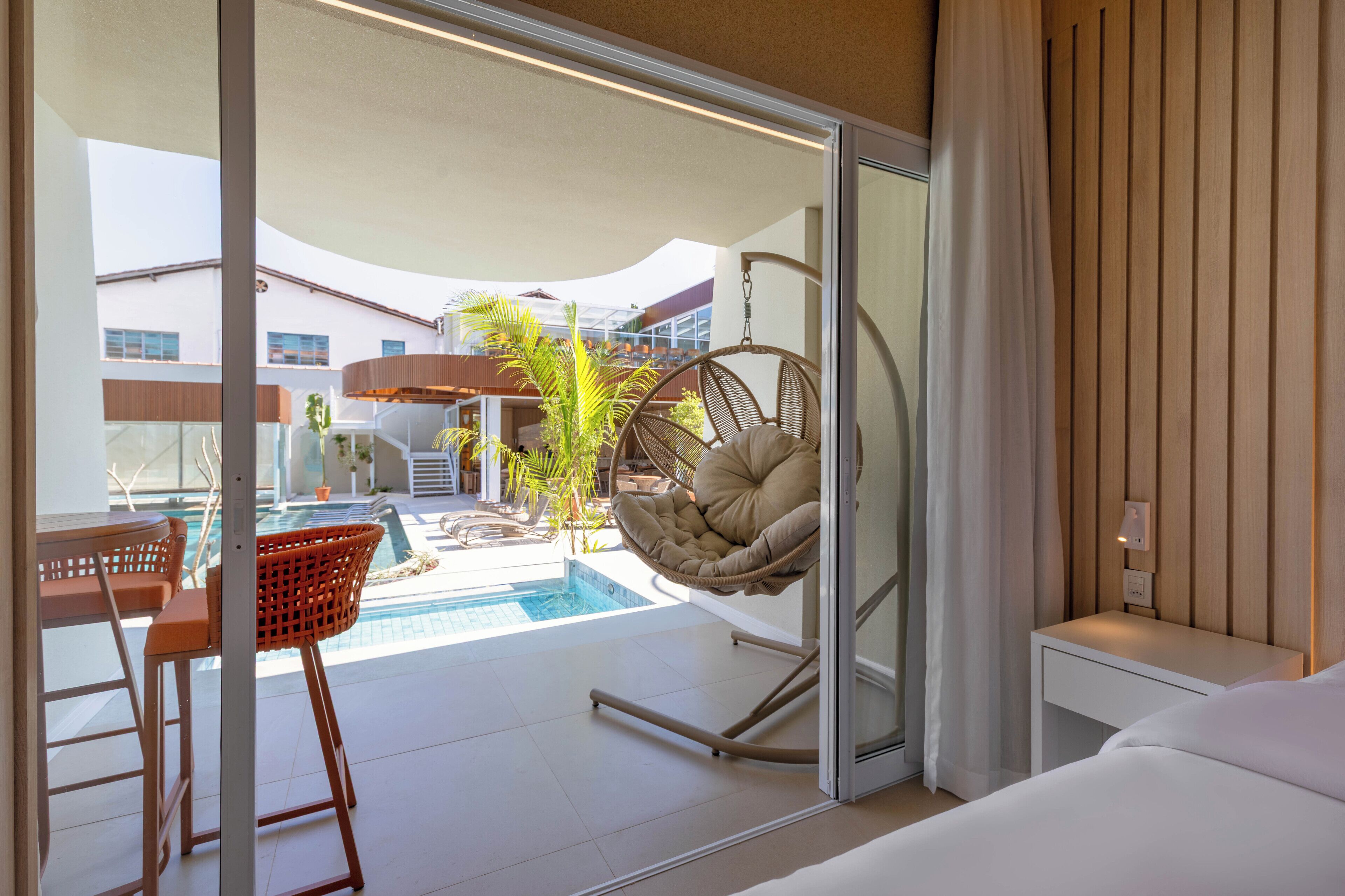 Camera d'albergo moderna con una porta vetrata che dà su una zona piscina, una sedia amaca e un tavolino con sedie.