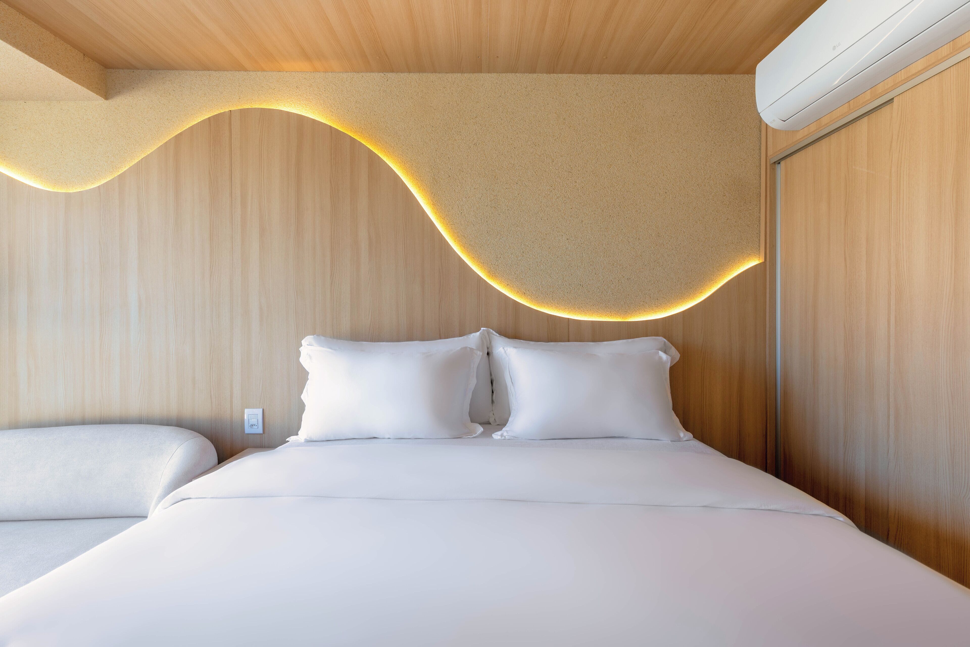Quarto moderno com teto de madeira, iluminação curva e cama com roupa de cama branca.