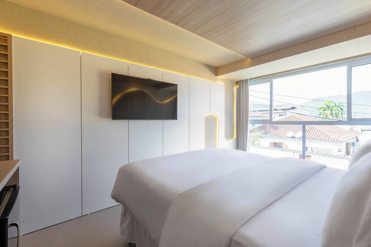 A modern bedroom with a large bed, a flat-screen TV mounted on the wall, and a view of the outside through large windows.