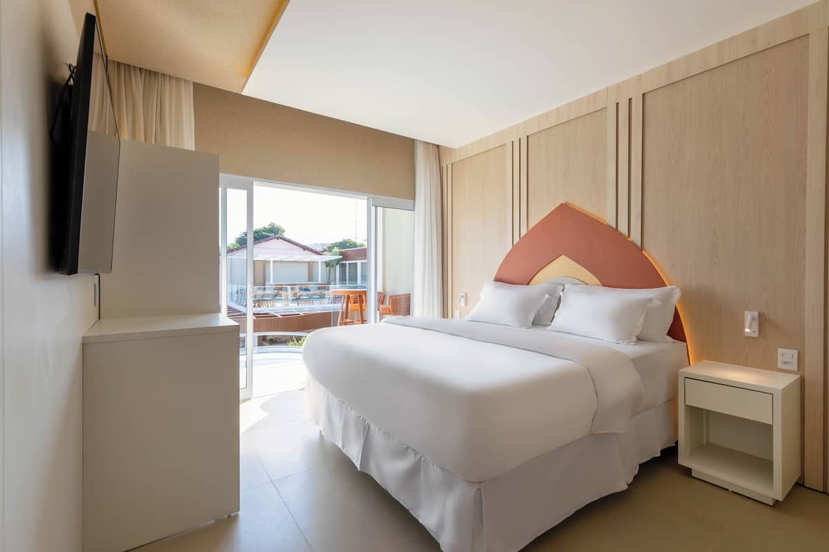 A hotel room with a large bed, a small bedside table, a TV mounted on the wall, and a sliding glass door leading to a balcony.
