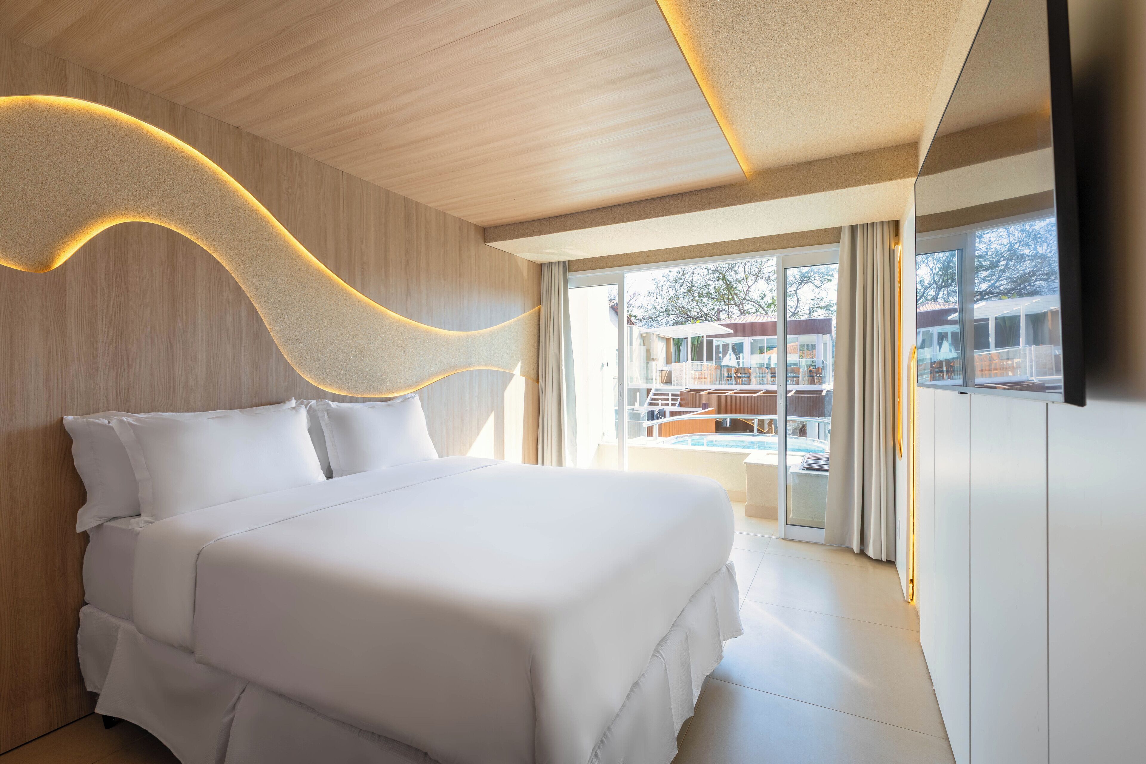A modern hotel room with a large bed, a wooden ceiling, and a view of the pool and outdoor area.