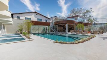 A modern pool area with a glass-enclosed upper deck, a clear blue pool, and a white building in the background.