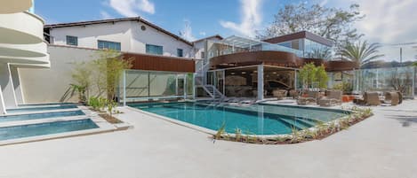 A modern pool area with a glass-enclosed upper deck, a clear blue pool, and a white building in the background.