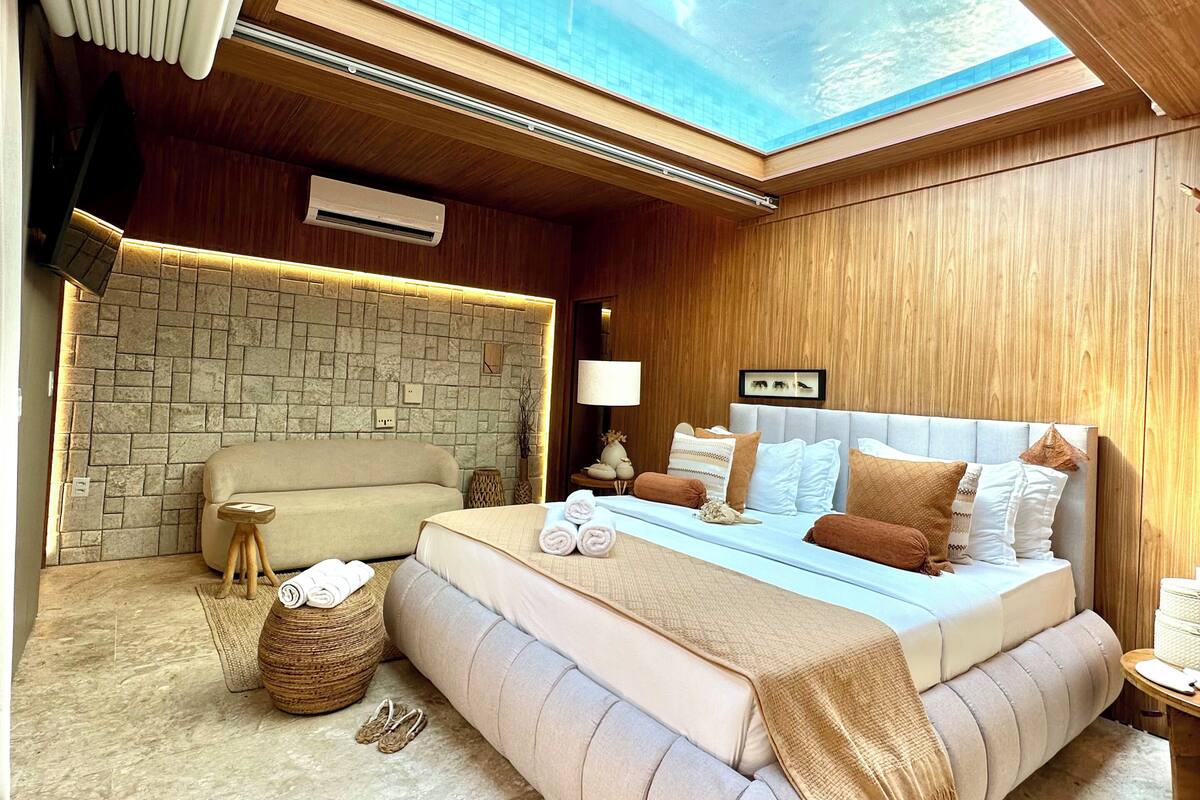 A bedroom with a skylight, a stone wall, a bed with pillows, and a small wooden stool.