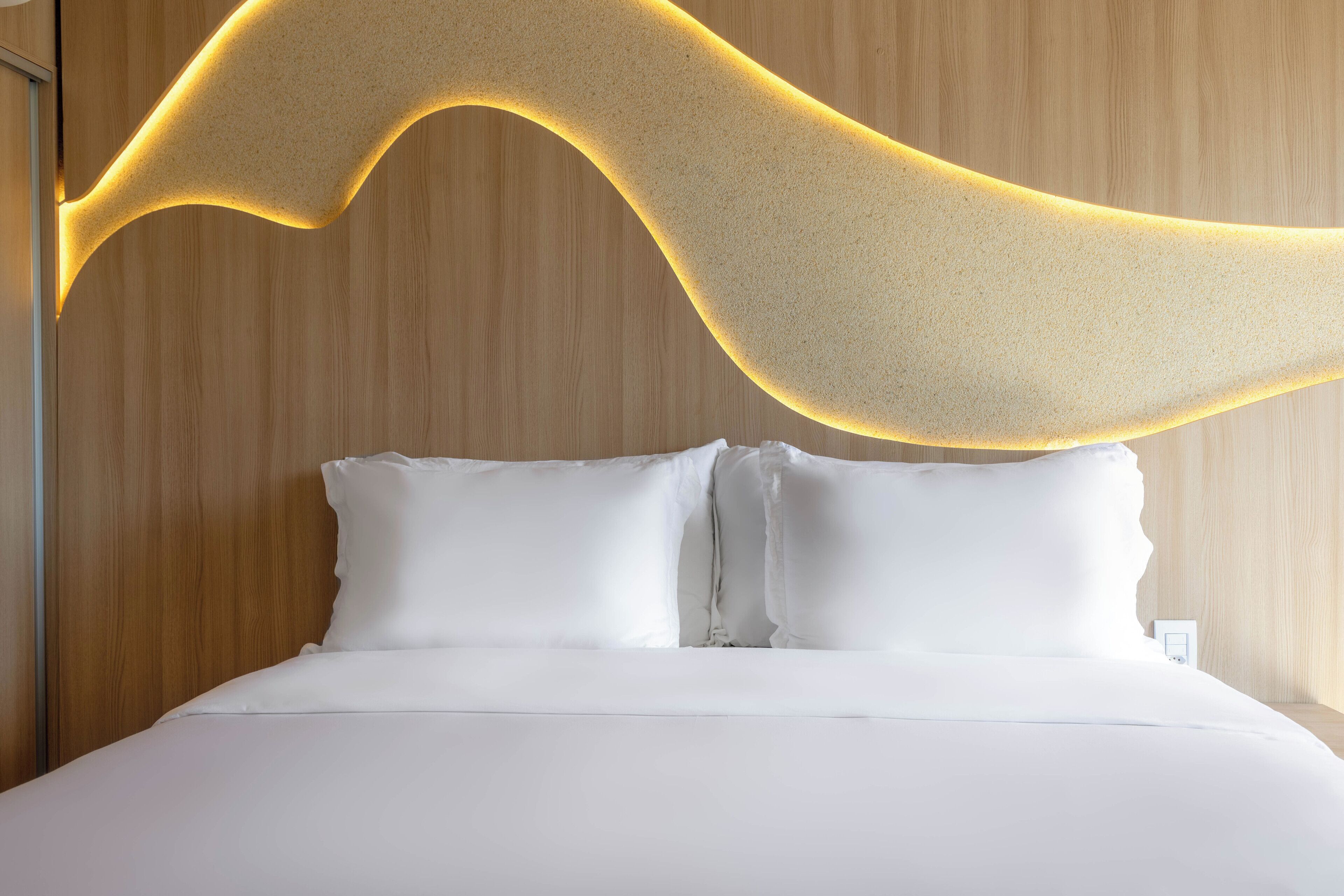 Une chambre d’hôtel moderne avec une tête de lit en bois, une literie blanche et un luminaire original placé au-dessus du lit.