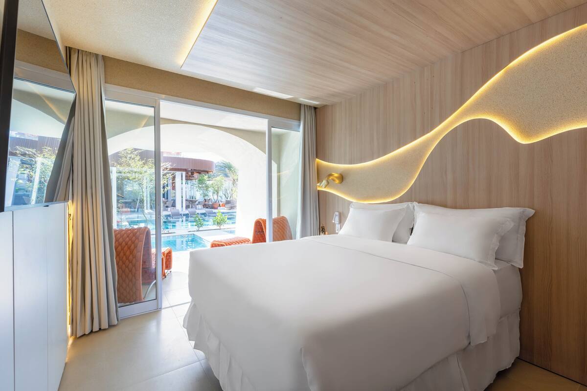 A modern hotel room with a large bed, a view of a swimming pool, and a wooden ceiling.
