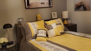 Un dormitorio con cama, mesita de noche, lámpara, marco de foto, un florero con flores y un cuadro en la pared.