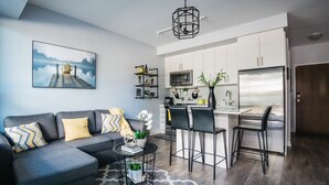 Una sala de estar moderna con un sofá gris, una mesa de centro y una zona de cocina con armarios blancos y un refrigerador de acero inoxidable.