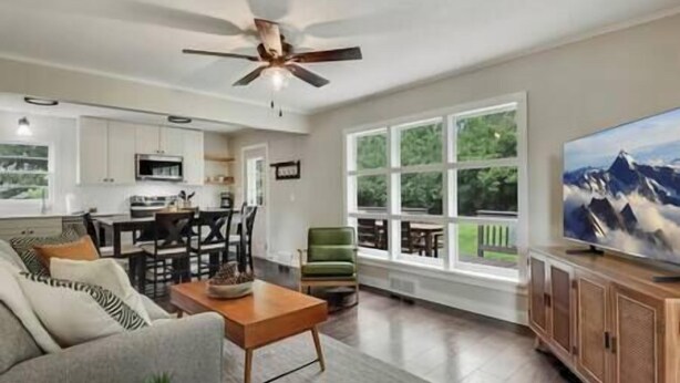 A spacious living area with a sofa, coffee table, and a television. A ceiling fan is visible, and there are large windows offering a view of greenery outside.