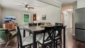 A dining area with a large table and chairs, a ceiling fan, and a refrigerator in the background.
