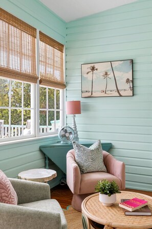 Une pièce avec un mur de couleur turquoise, un tableau sur le thème de la plage, un ventilateur, une chaise rose et une petite table sur laquelle se trouvent une plante et des livres.