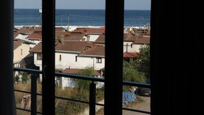 A view through a window with a balcony overlooking a coastal area with buildings and the sea. - Koç Guest House (Demirkoy)