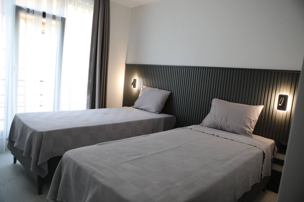 Habitación de hotel con dos camas individuales, un cabecero moderno y una ventana con cortinas.