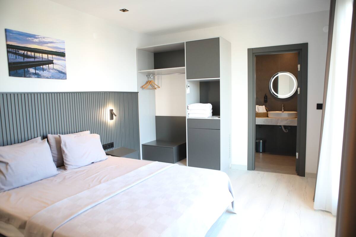 Un dormitorio moderno con cama, mesitas de noche, armario, espejo y baño con lavamanos e inodoro.
