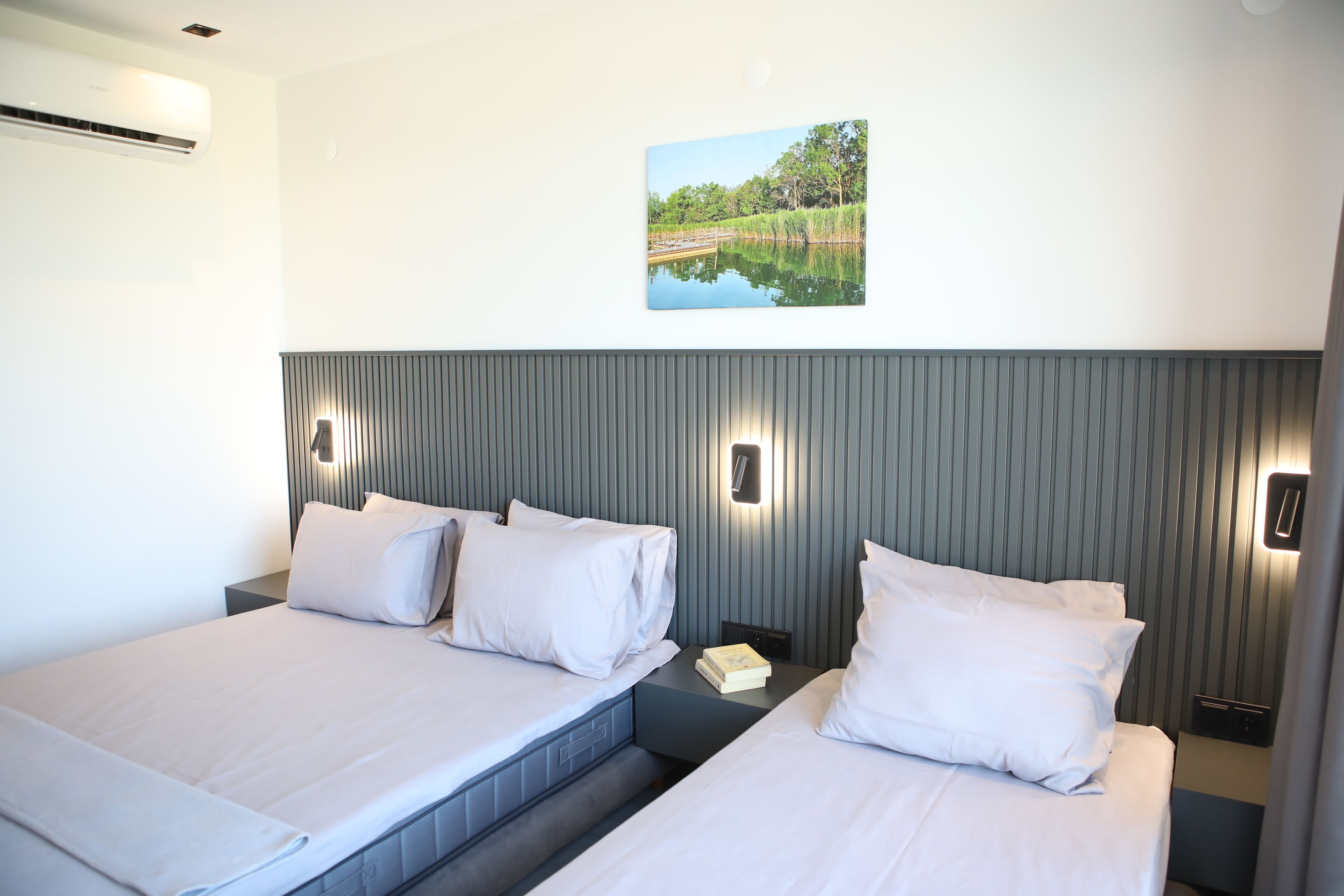 A hotel room with two beds, a wall-mounted air conditioner, a nightstand with a book, and a framed picture of a lake.