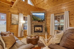 A log cabin interior with a stone fireplace, leather sofas, a flat-screen TV, and a large window. - Gatlinburg Getaway - Downtown, Hot Tub, Theatre (Gatlinburg)
