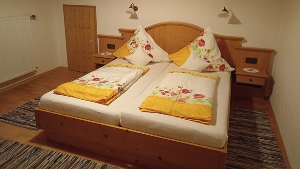 Cama doble con ropa de cama floral, cabecera de madera, dos mesitas de noche y un armario de madera.