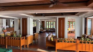 Eine geräumige Lobby mit Holzmöbeln, einem Deckenventilator und großen Fenstern, die natürliches Licht hereinlassen.