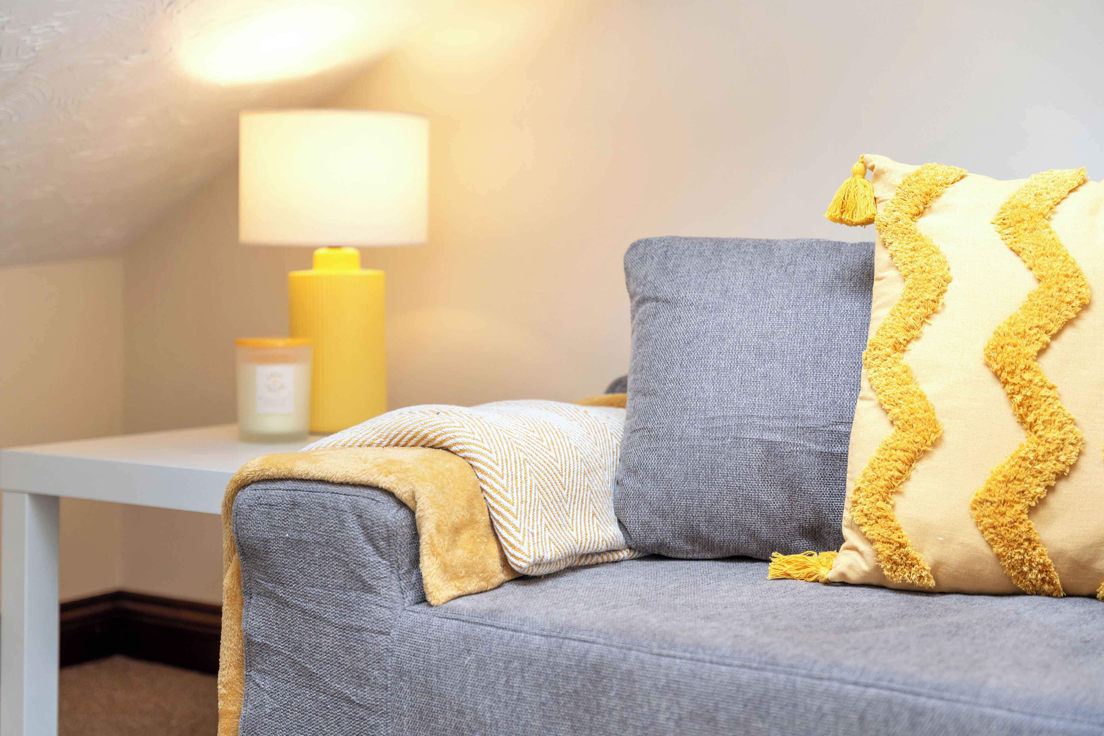 Une pièce chaleureuse avec un canapé gris, une lampe jaune et une petite table sur laquelle est posée une bougie.