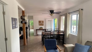 A spacious living room with a wooden floor, a ceiling fan, a dining area with a table and chairs, and large windows offering a view of the outdoors.