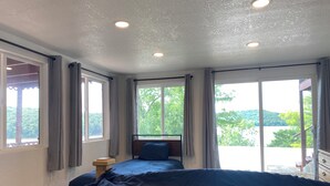 A bedroom with a large bed, two windows, and a view of greenery and water.