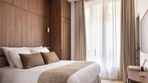 Una habitación de hotel moderna con una cama grande, paneles de madera y vistas a la ciudad a través de cortinas translúcidas.