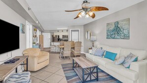 A living room with a TV, sofa, and chairs, a ceiling fan, and a kitchen in the background.