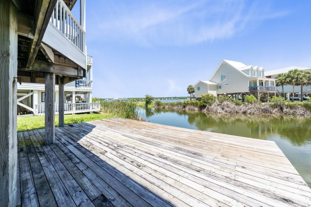 Una terraza de madera que conduce a una propiedad frente al agua, con un edificio blanco y un muelle que se adentra en una masa de agua tranquila.