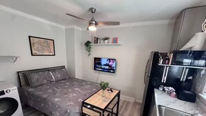 Habitación pequeña con cama, una mesita, televisor, ventilador de techo, microondas y lavamanos.