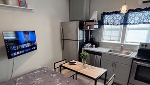 Cocina compacta con refrigerador, estufa y lavamanos. Un televisor de pantalla plana está montado en la pared, y hay una pequeña mesa con un florero en la zona de comedor.