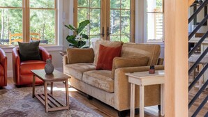 Ein geräumiges Wohnzimmer mit hoher Decke, großen Fenstern, einer Holztreppe und einer gemütlichen Sitzecke mit Sofa und Sesseln.