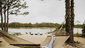 Eine Hängematte, zwischen zwei Bäumen an einem sandigen Strand befestigt, mit einem See und Picknicktischen im Hintergrund.