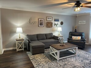 A living room with a sectional sofa, two side tables with lamps, a coffee table, a black leather chair, and a ceiling fan. - Charming 2-bedroom corporate townhome in pleasant Madison. (Madison)