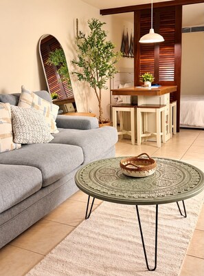 Een woonkamer met een grijze bank, een ronde salontafel met een geweven mand, en een eethoek met een houten tafel en stoelen.