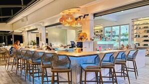 Een moderne bar met een lange toonbank, rieten barstoelen en diverse flessen en glazen.