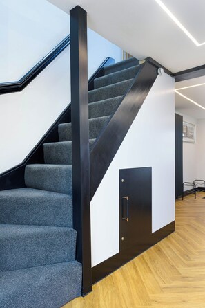 Un escalier avec des marches recouvertes d’une moquette grise et des rampes noires, menant à un palier où se trouve une porte.