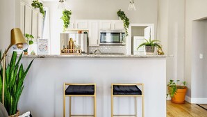 A kitchen with white cabinets, a stainless steel oven, a microwave, and a hanging plant. There are two bar stools with black cushions. - Short North & Downtown & OSU | Near High Street | Free Parking | 5,800+ Reviews (Columbus)