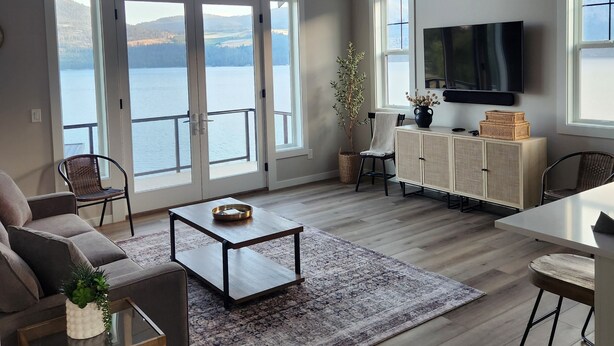 A spacious living room with a large window overlooking a lake, a ceiling fan, and a flat-screen TV mounted on the wall. - Lake Chelan view home 1-bedroom in Manson with AC (Manson)