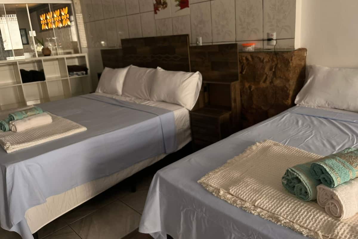 Habitación de hotel con dos camas, cada una con cubrecama y almohadas blancas. Incluye una pequeña mesa de madera y un estante con artículos de aseo.
